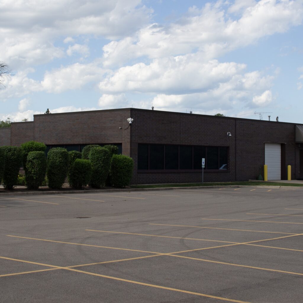 Large brown brick building with dark windows, closed door, and adjacent asphalt parking lot.