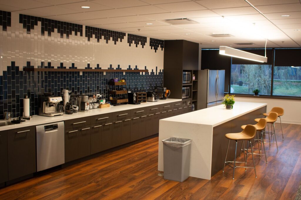 Modern kitchenette with dark cabinets, white countertop, and large center island with barstools. Featuring kitchen appliances, dark blue tile backsplash, and a large window.