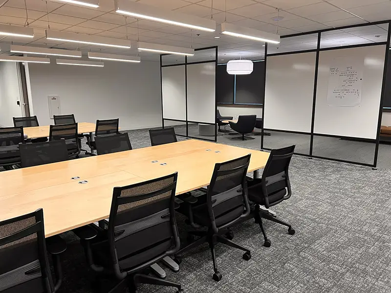 Office meeting room with a large conference table and numerous black office chairs, separated by a glass partition from a lounge area.