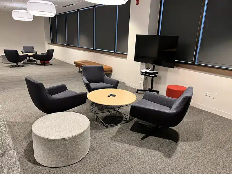 Modern office area with gray armchairs, round wooden table, white ottoman, and TV on a stand.